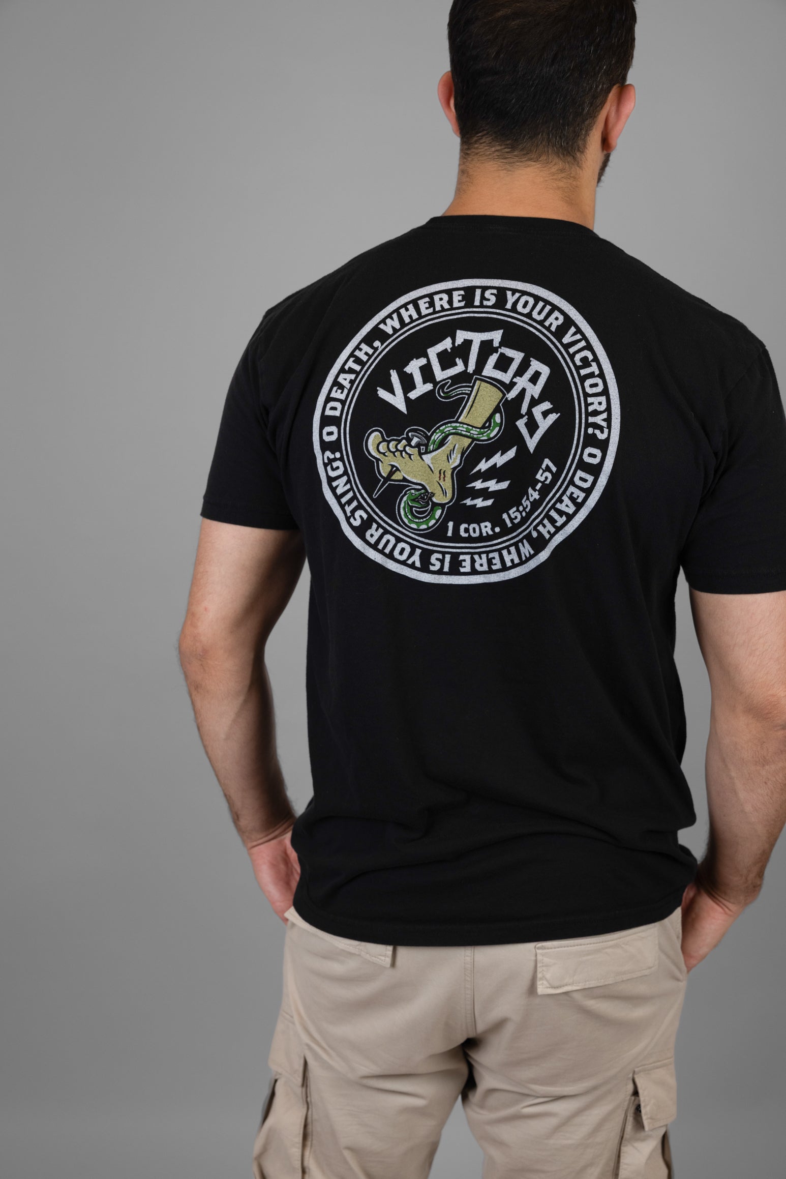 Person wearing a black t-shirt with a VICTORY logo and text on the back, standing against a gray background