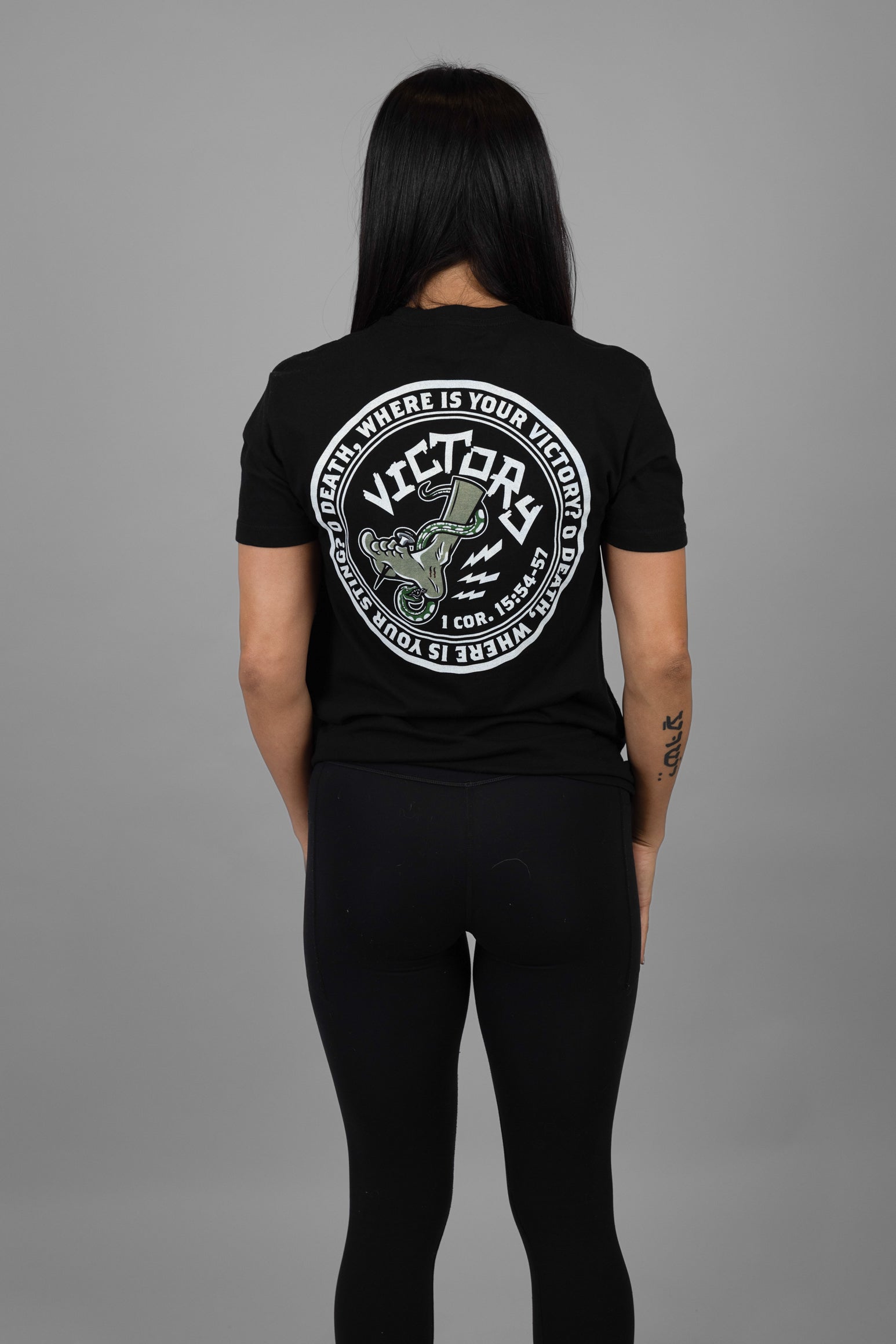 Person wearing a black t-shirt with a VICTORY logo and text on the back, standing against a gray background