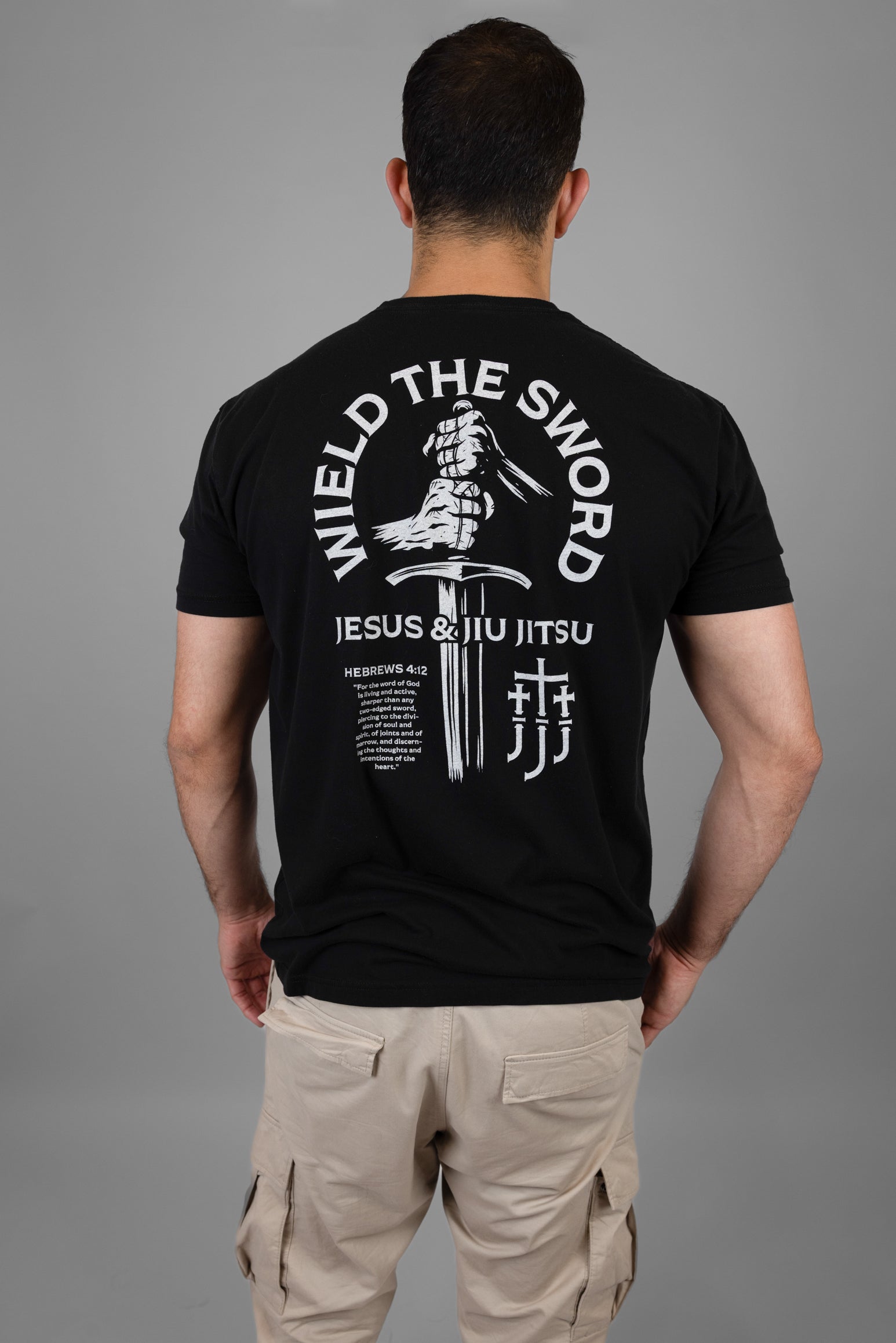 Person wearing a black t-shirt with 'Wield the Sword Jesus & Jiu Jitsu' design on the back, standing against a gray background.