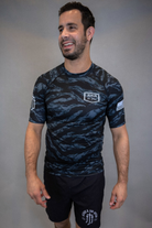 man wearing camo rashguard with Jesus and Jiu Jitsu logo on left chest and flag on left sleeve, and JJJ logo on right sleeve