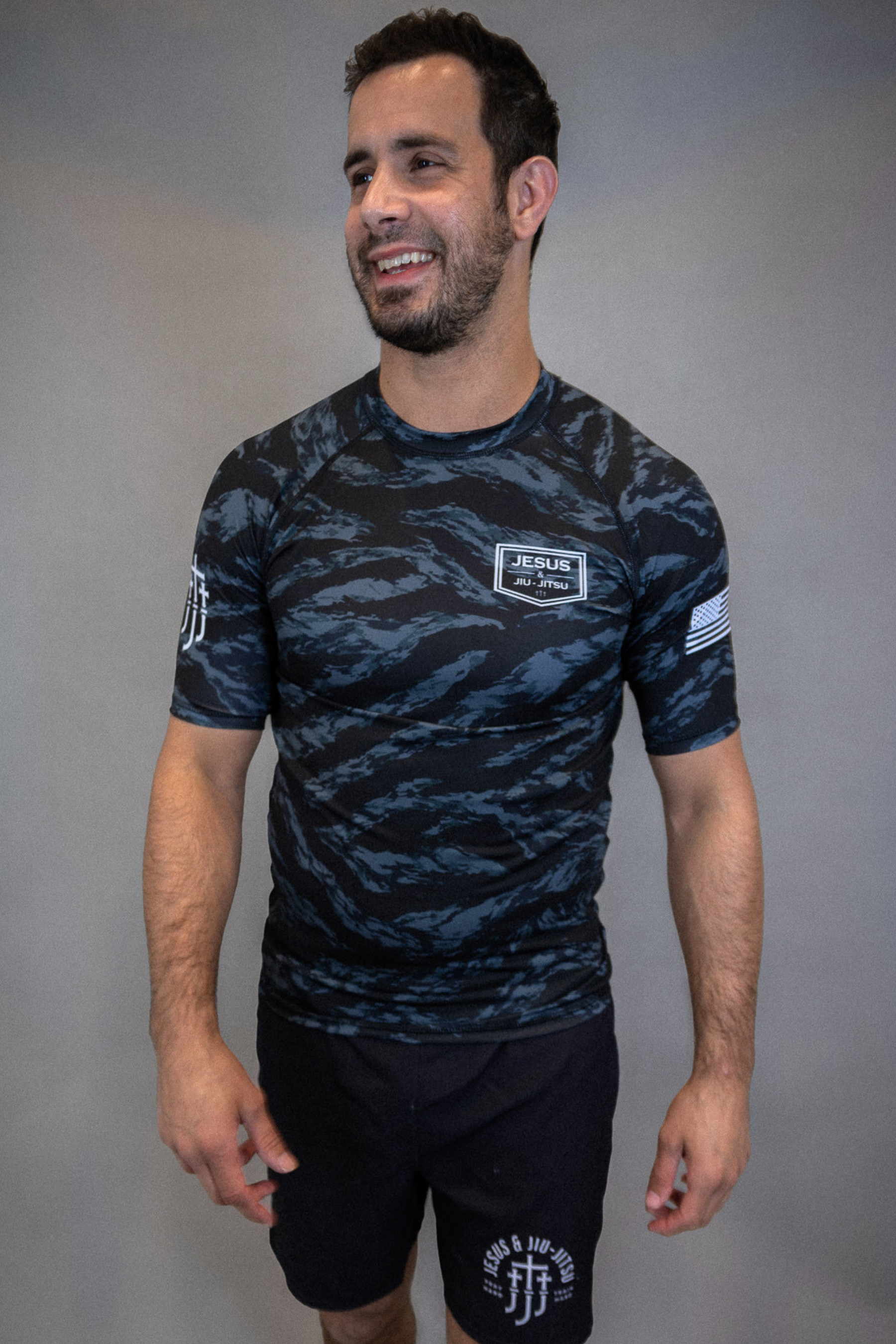 man wearing camo rashguard with Jesus and Jiu Jitsu logo on left chest and flag on left sleeve, and JJJ logo on right sleeve