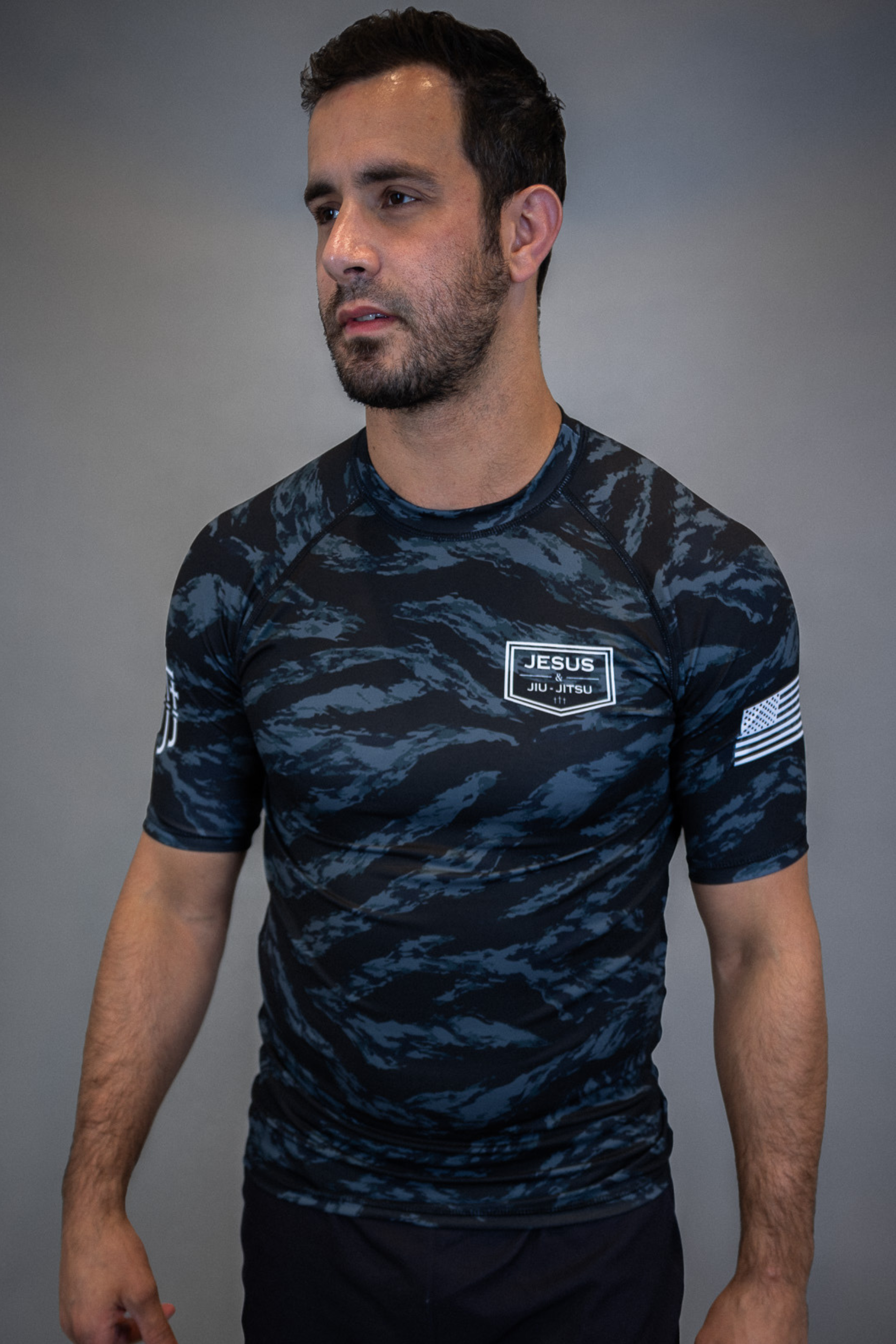 man wearing camo rashguard with Jesus and Jiu Jitsu logo on left chest and flag on left sleeve