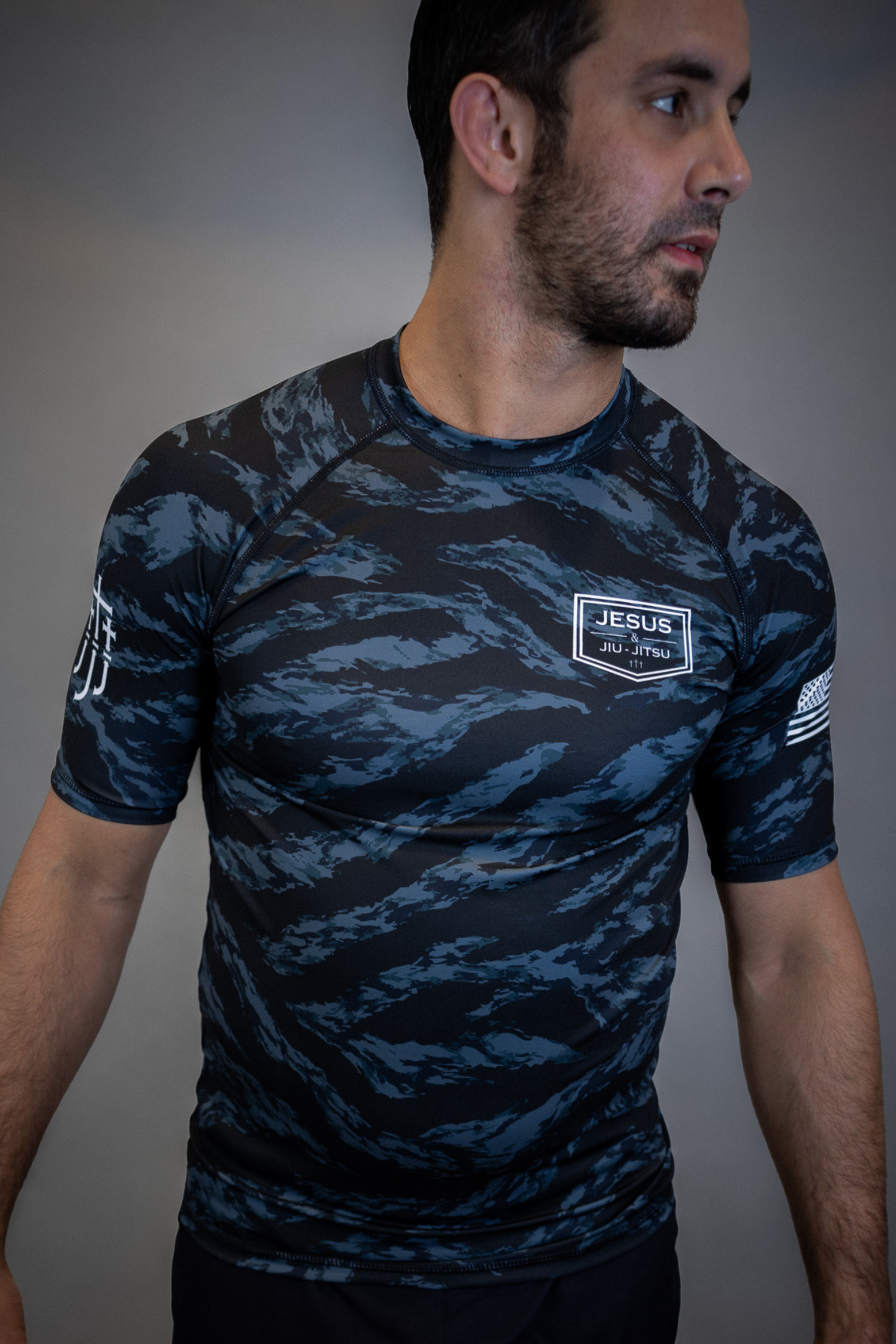 man wearing camo rashguard with Jesus and Jiu Jitsu logo on left chest and JJJ on right sleeve and flag on left sleeve
