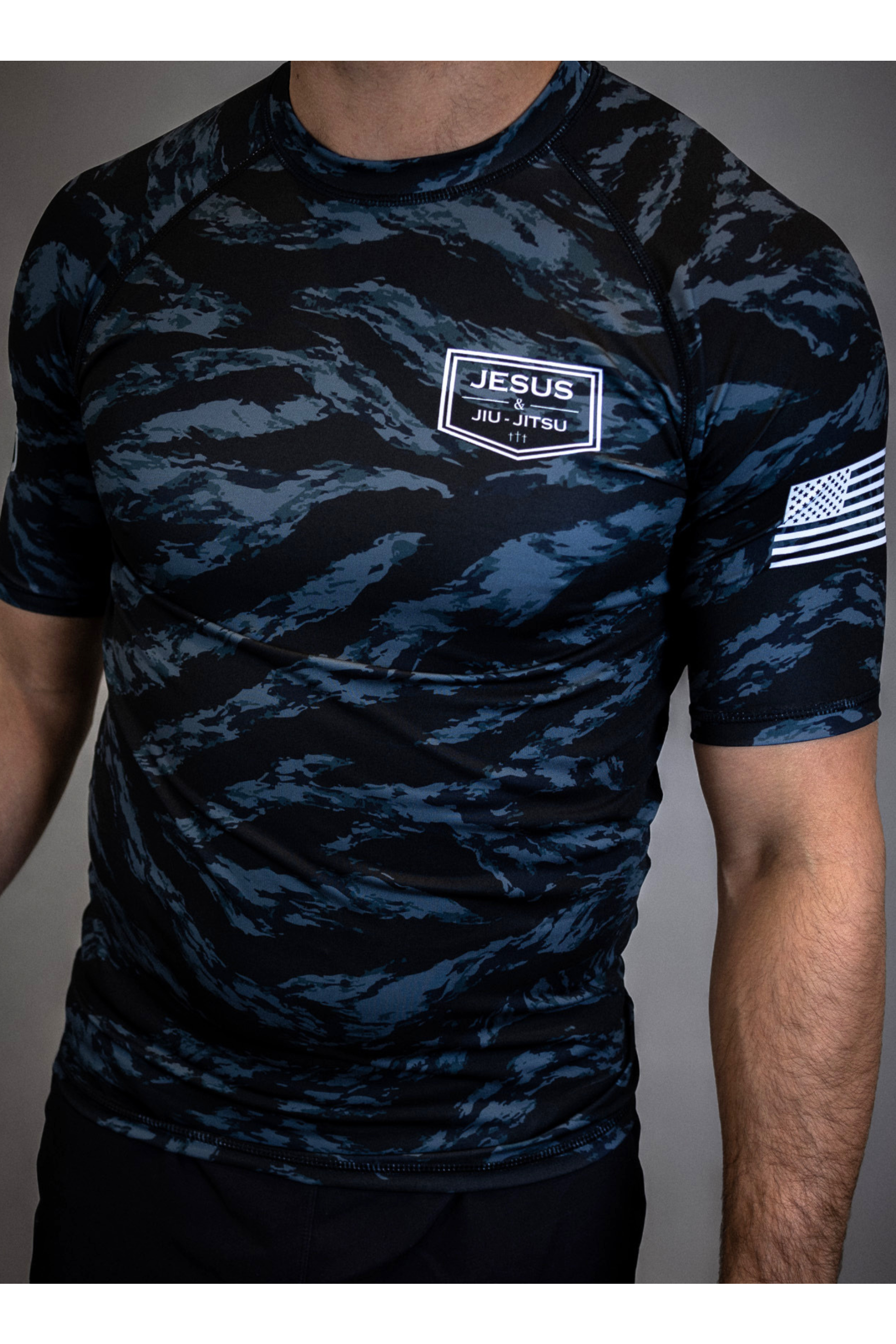 man wearing camo rashguard with Jesus and Jiu Jitsu logo on left chest and flag on left sleeve