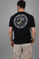 Person wearing a black t-shirt with a  VICTORY logo and text on the back, standing against a gray background