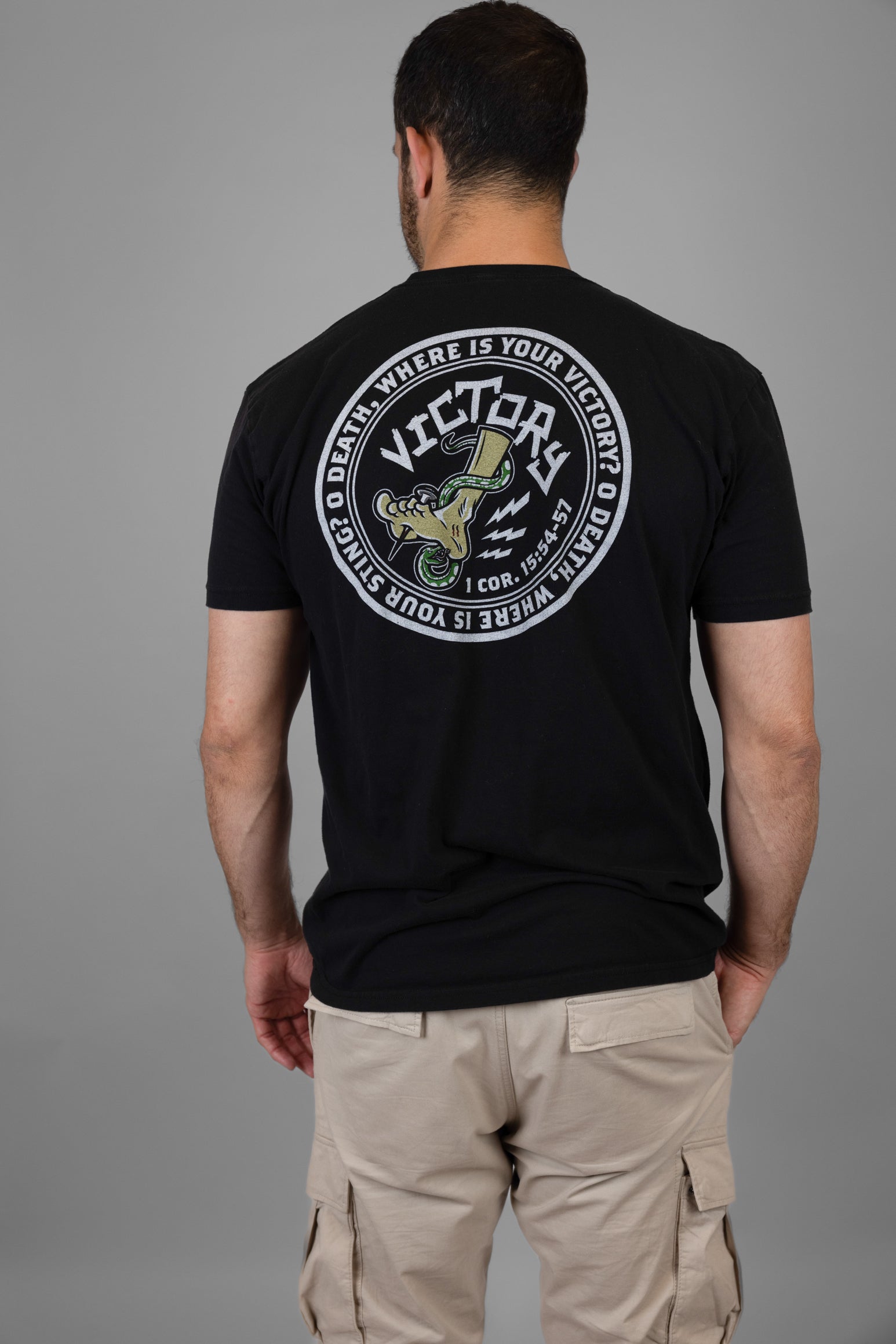 Person wearing a black t-shirt with a  VICTORY logo and text on the back, standing against a gray background