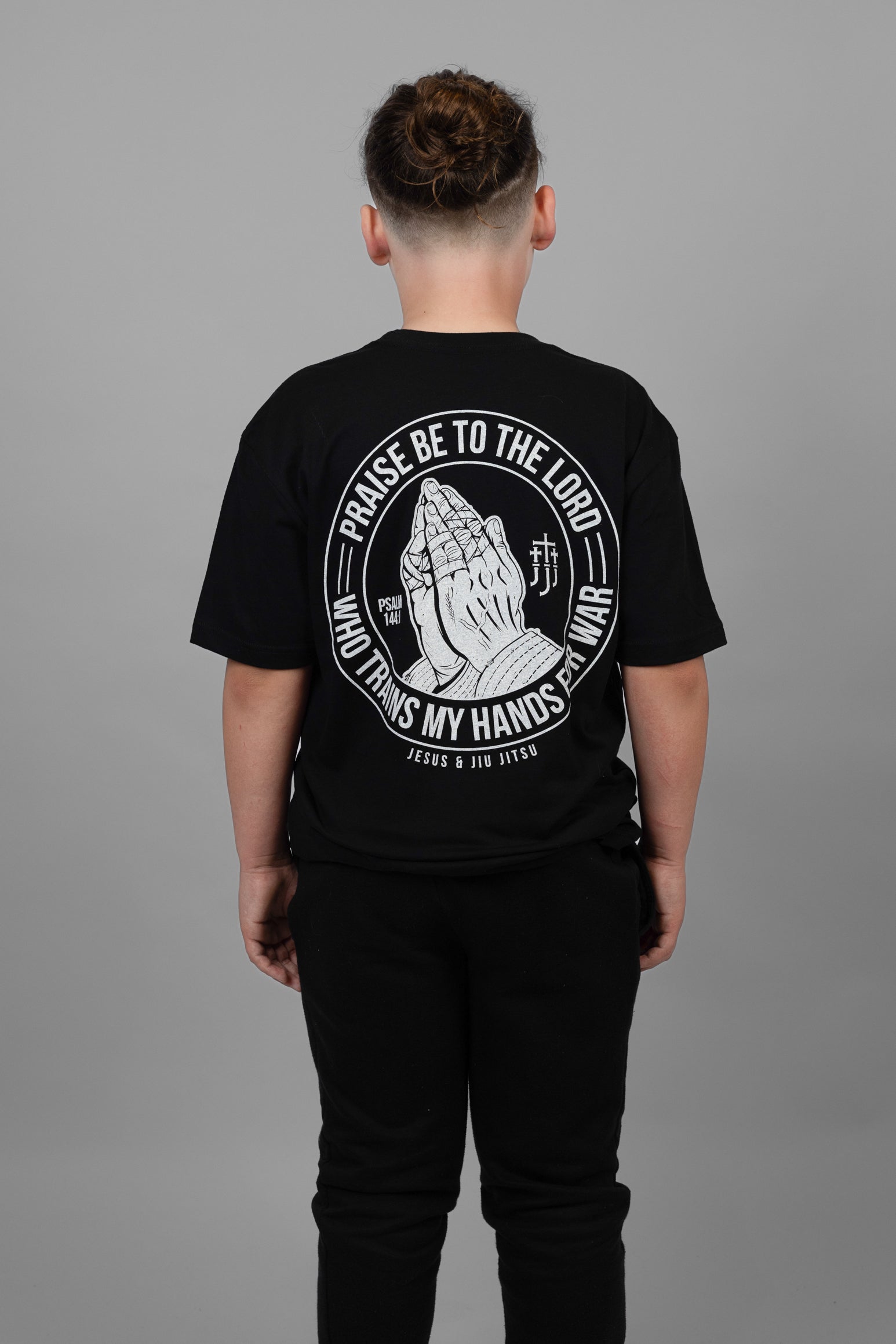 Child wearing black tee with praying hands graphic on back with text "PRAISE BE TO THE LORD WHO TRAINS MY HANDS FOR WAR"