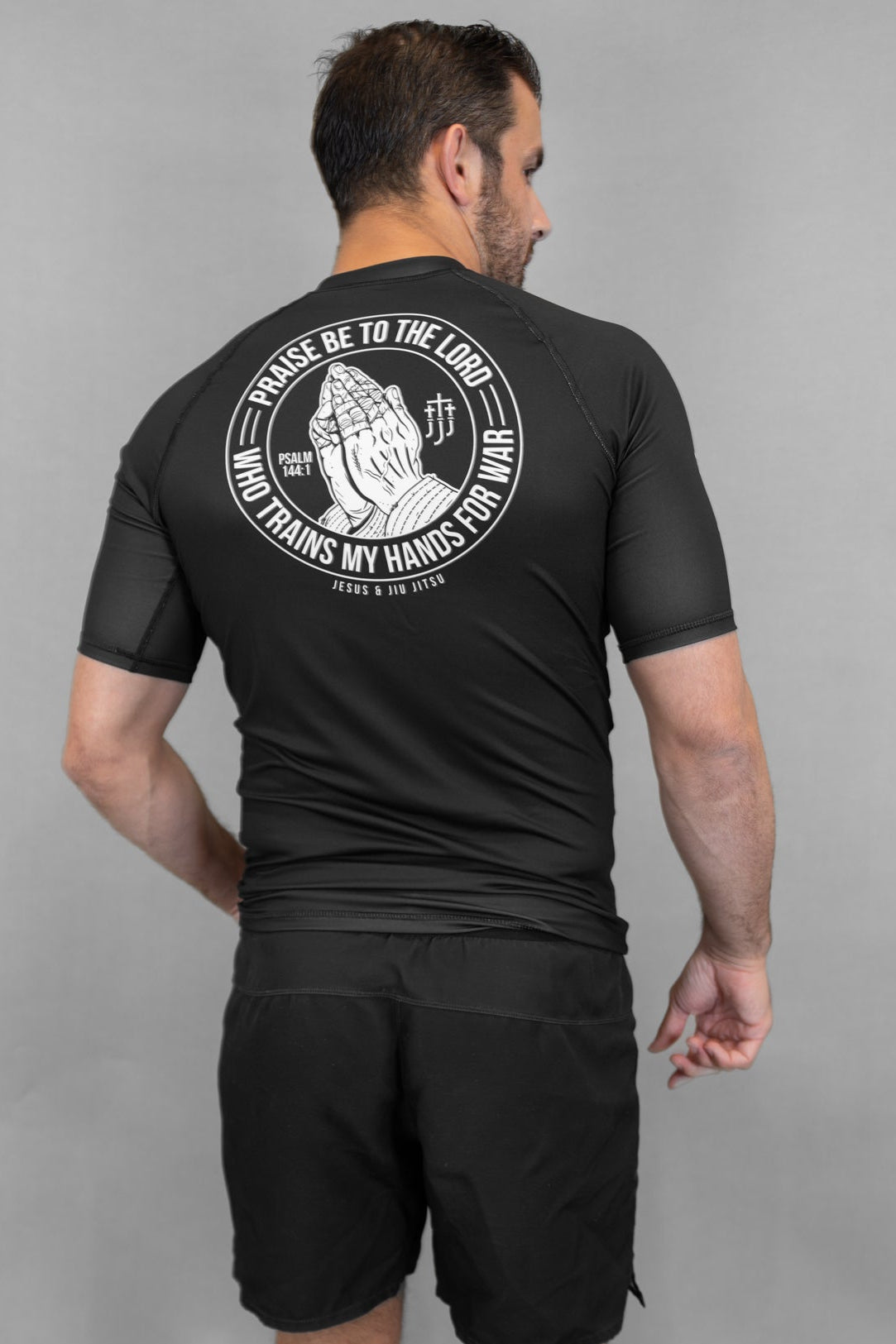 Person wearing a black Prayer hands rashguard and shorts with a logo on the back against a gray background