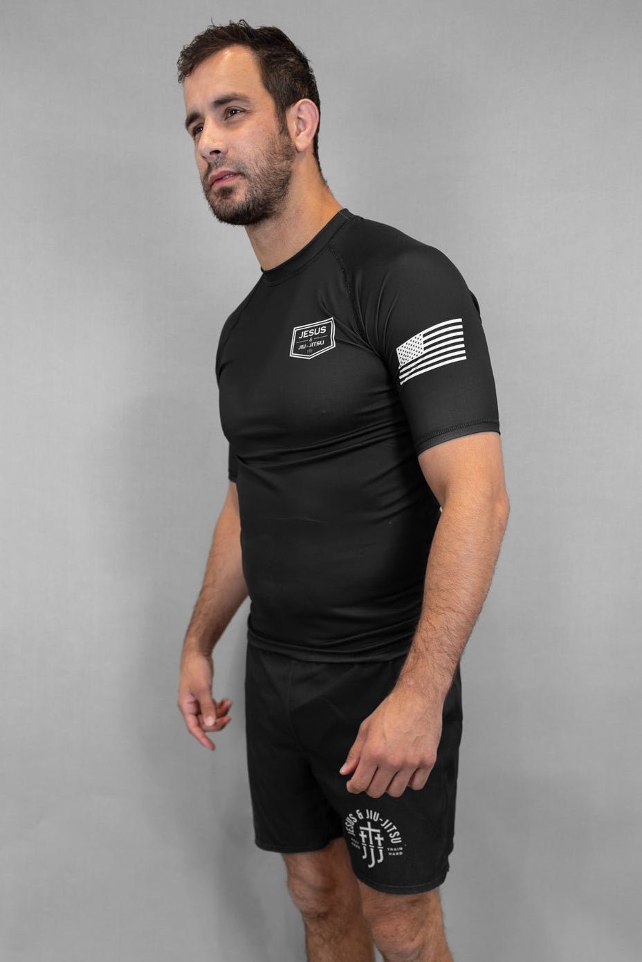 Man wearing a black Rashguard with JJJ logo on chest and flag on sleeve on a gray background