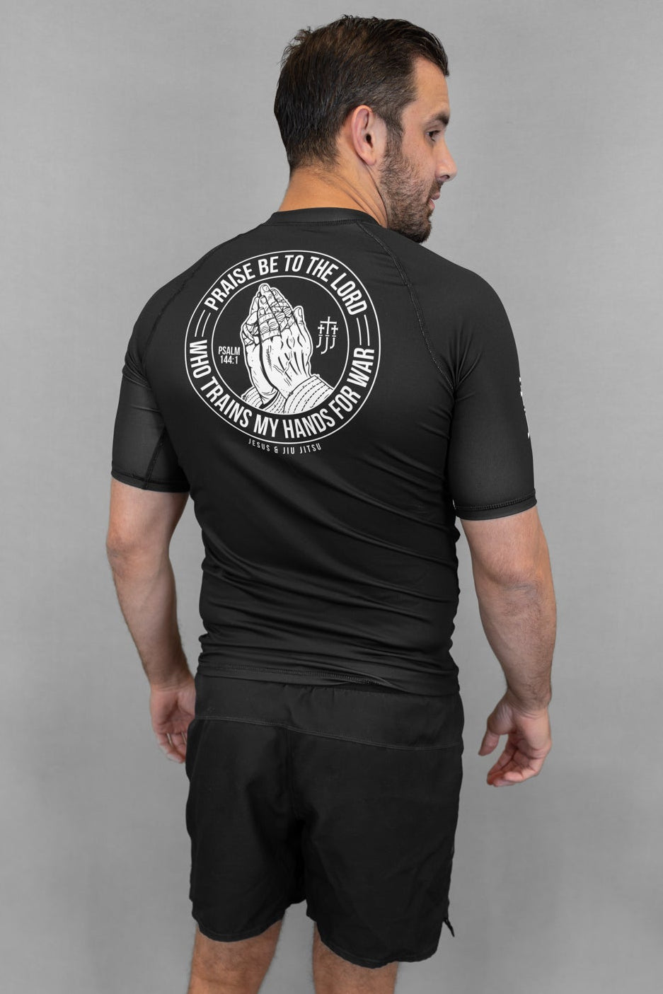 Person wearing a black Prayer hands rashguard and shorts with a logo on the back against a gray background
