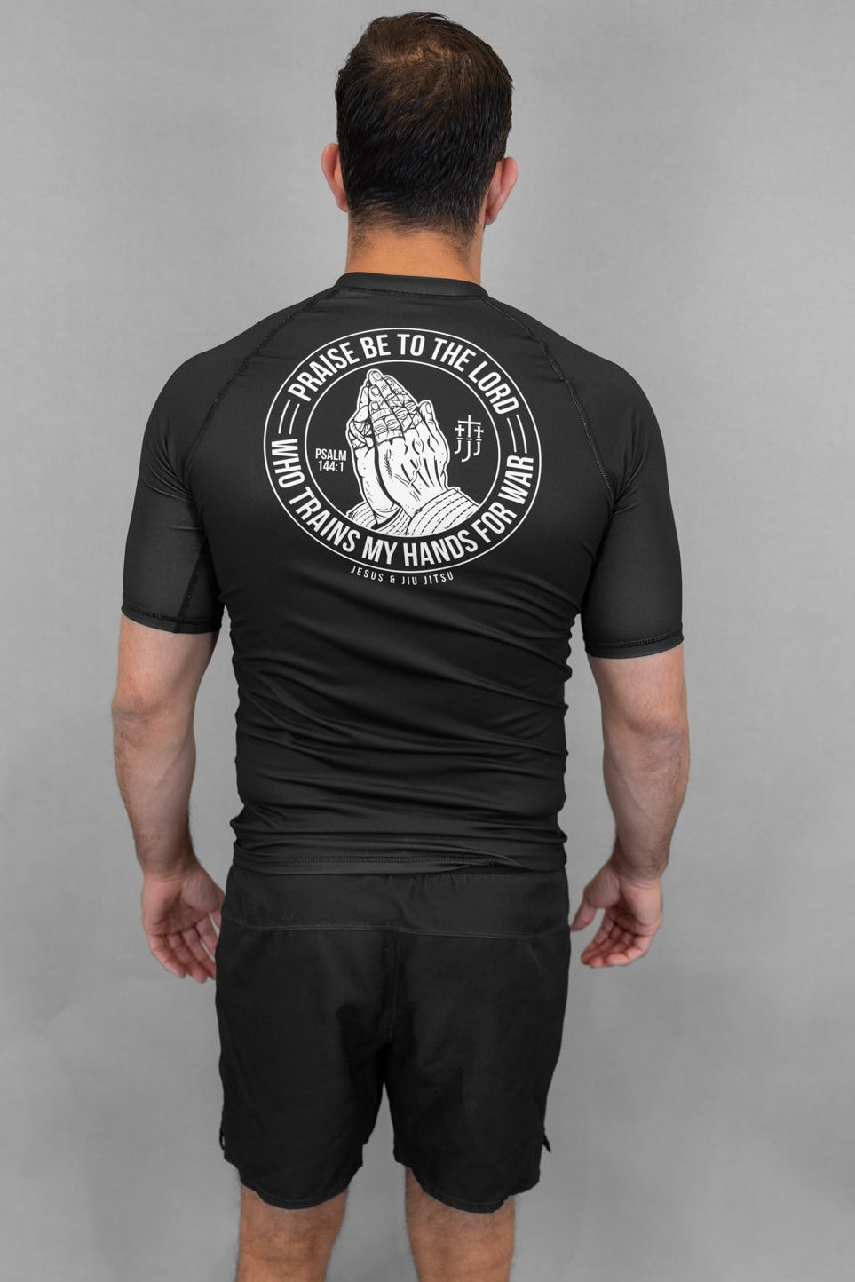 Person wearing a black Prayer hands rashguard and shorts with a logo on the back against a gray background