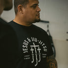 Man wearing a black t-shirt with white "JESUS & JIU-JITSU" text and logo on a gray background