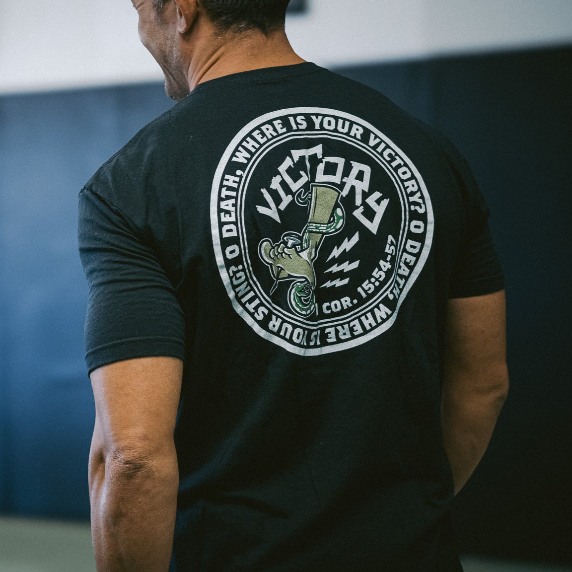 Person wearing a black t-shirt with a  VICTORY logo and text on the back, standing against a gray background